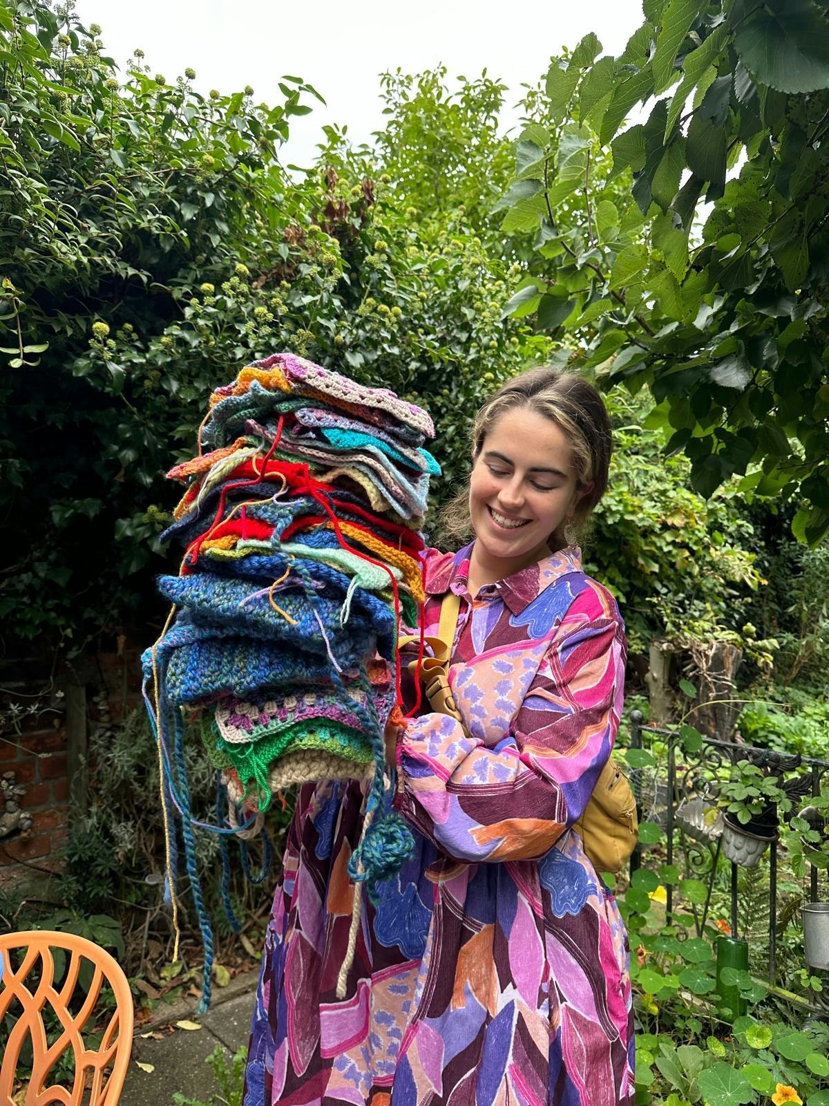 A woman in a patterned dress smiling while holding a tall pile of colourful crocheted and knitted squares in a garden